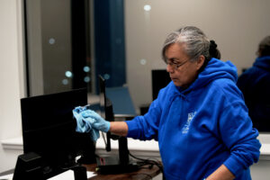 Woman in blue sweatshirt and blue gloves wiping a computer screen with a blue towel